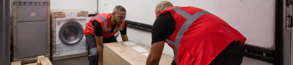 Deux hommes en gilet de sécurité rouge déplacent un gros carton dans un camion de livraison